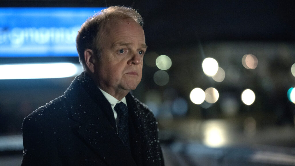 Ein Mann mittleren Alters in dunklem Mantel und Anzug steht bei Nacht im Schneefall. Im unscharfen Hintergrund sind Lichter einer Stadt oder Strasse zu sehen. Die Szene wirkt dramatisch und atmosphärisch.