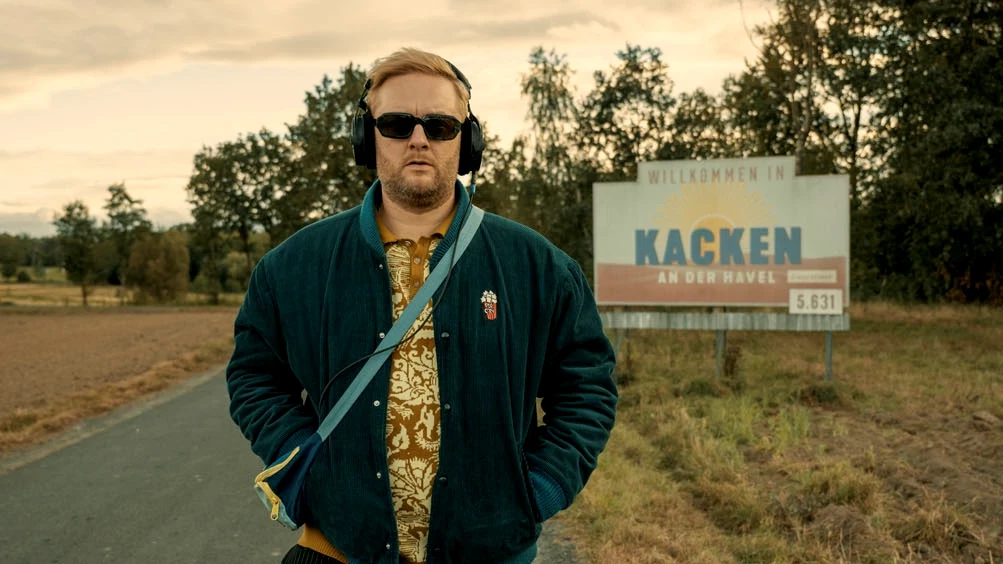 Ein Mann mit Sonnenbrille und Kopfhörern steht auf einer Landstrasse. Er trägt eine grüne Jacke und ein gemustertes Hemd. Im Hintergrund ist ein Ortsschild mit der Aufschrift «Willkommen in Kacken an der Havel» zu sehen, umgeben von Feldern und Bäumen.