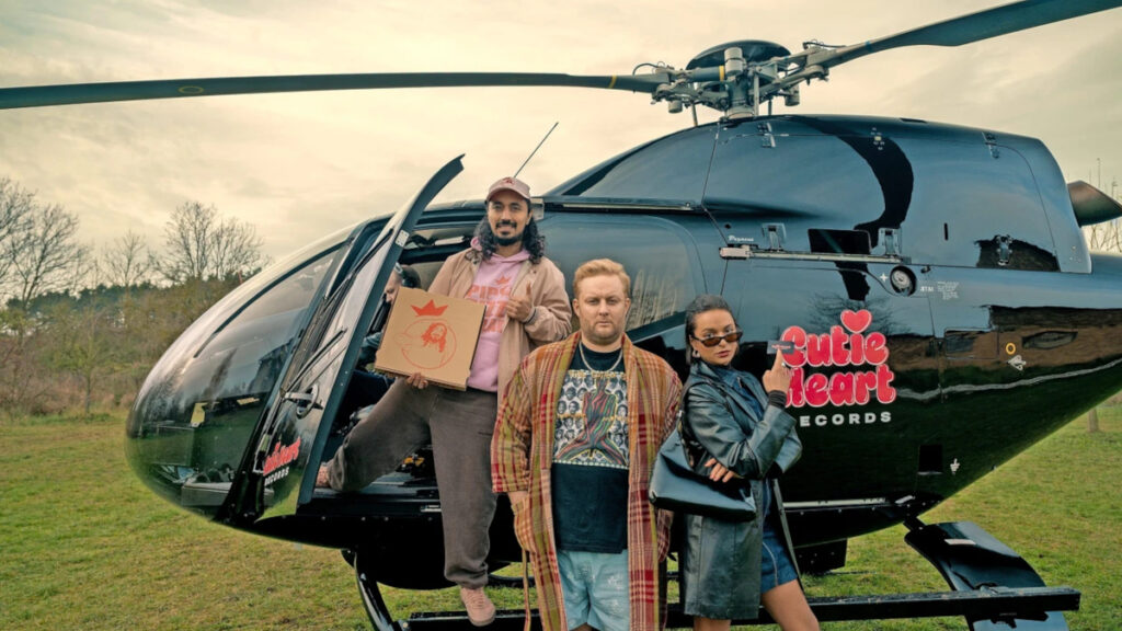 Drei Personen stehen vor einem schwarzen Helikopter auf einer Wiese. Links ein Mann mit Kappe und rosa Pullover, der eine Schachtel hält. In der Mitte ein Mann mit kariertem Mantel, rechts eine Person in blauer Jacke und Sonnenbrille. Der Helikopter trägt das Logo «Cutie Heart Records».