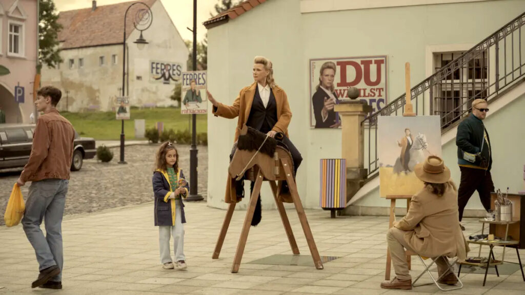 Eine Frau sitzt auf einem Sattel auf einem Holzgestell. Davor ein Maler, der sie auf einem Pferd malt. Umstehende Personen beobachten die Szene. Im Hintergrund sind alte Gebäude und Plakate zu sehen. 