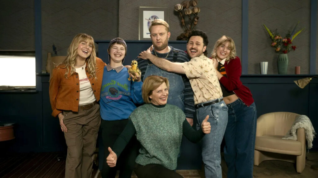 Sechs Personen posieren fröhlich in einem modernen Wohnzimmer. Sie tragen lässige Kleidung und umarmen sich, während sie in die Kamera lächeln. Das Set wirkt wie aus einer Comedy-Serie mit gemütlicher Einrichtung und warmer Beleuchtung.