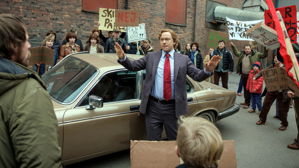 Ein Mann in dunklem Anzug und roter Krawatte steht vor einem beigen Auto und gestikuliert zu einer Menschenmenge mit Protestschildern. Die Szene spielt vor Backsteingebäuden und wirkt wie aus einer TV-Serie der 1980er Jahre.