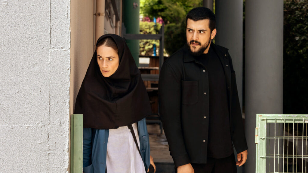 Eine Frau mit schwarzem Kopftuch und ein Mann mit dunklem Bart stehen nebeneinander vor einem Gebäude. Beide tragen dunkle Kleidung und blicken zur Seite. Im Hintergrund sind weisse Wände und grüne Pflanzen zu sehen.