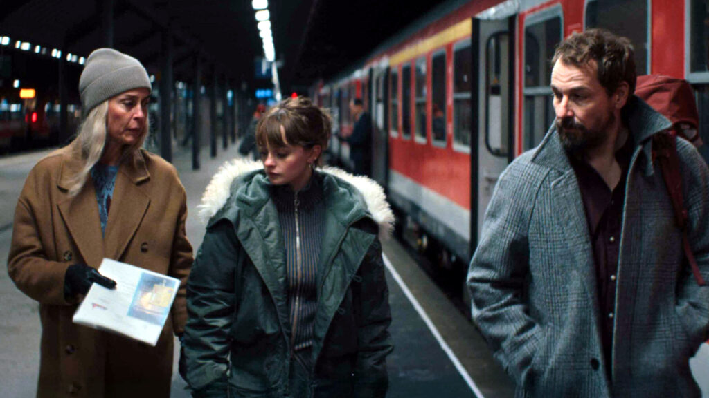 Drei Personen stehen nachts auf einem Bahnsteig vor einem roten Zug. Eine Frau in braunem Mantel und Mütze hält Papiere, daneben eine jüngere Frau in grüner Winterjacke und ein bärtiger Mann im grauen Pullover. Szene aus einer TV-Serie.