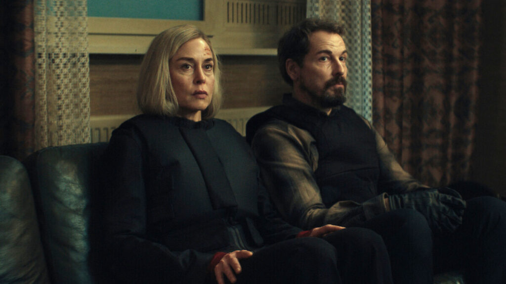 Eine blonde Frau und ein bärtiger Mann sitzen angespannt nebeneinander auf einem grünen Sofa. Beide tragen dunkle Kleidung und blicken ernst zur Seite. Im Hintergrund sind gemusterte Vorhänge und eine vintage Inneneinrichtung zu sehen.