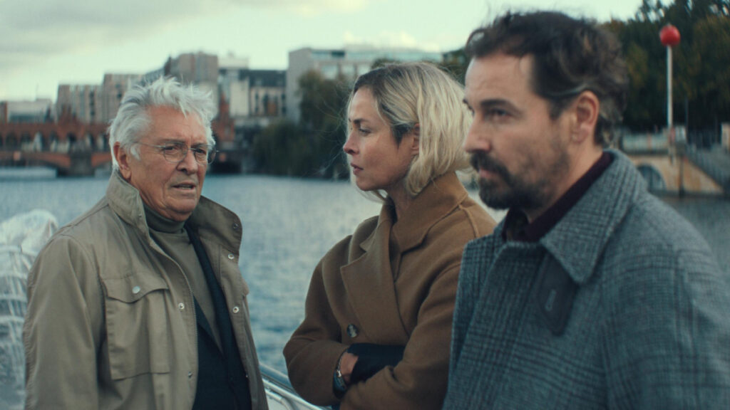 Drei Personen stehen am Ufer eines Gewässers: ein älterer Mann mit grauer Jacke links, eine blonde Frau in braunem Mantel in der Mitte und ein bärtiger Mann in grauem Pullover rechts. Im Hintergrund sind Gebäude und eine Brücke zu sehen.