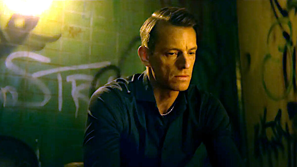 Ein Mann in dunkler Jacke sitzt in gedimmtem, gelblich-grünem Licht. Sein Gesicht ist nachdenklich, im Hintergrund sind unscharfe Graffiti an einer Wand zu sehen. Die atmosphärische Beleuchtung verleiht der Szene eine düstere, cineastische Stimmung.