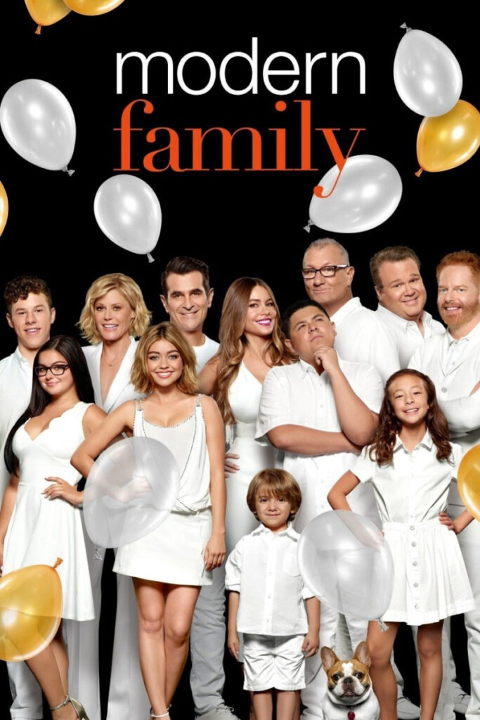 Cast-Foto der TV-Serie «Modern Family»: Mehrere Erwachsene und Kinder in weisser Kleidung posieren vor schwarzem Hintergrund mit dem Serientitel. Weisse und goldene Ballons schmücken das Bild. Ein kleiner Hund ist ebenfalls zu sehen.