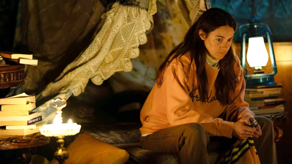 Eine junge Frau in orangem Pullover sitzt bei Kerzenschein auf einem Sessel. Um sie herum sind Bücher, eine Petroleumlampe und vintage Gegenstände arrangiert. Die warme Beleuchtung schafft eine intime Atmosphäre von Einsamkeit.