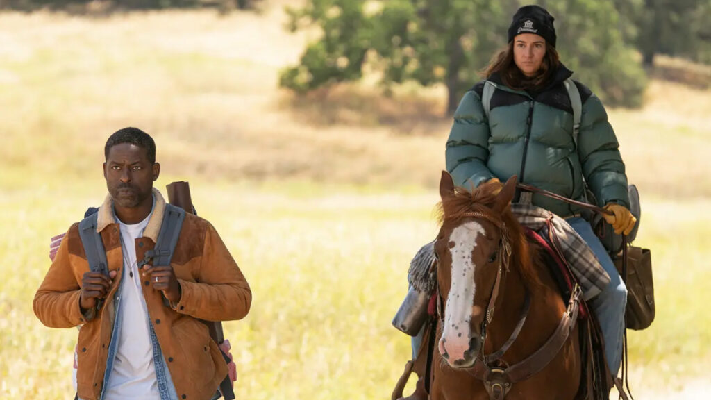 Ein Mann in brauner Jacke mit Rucksack geht zu Fuss durch eine sonnige Landschaft, während eine Frau in grüner Winterjacke und schwarzer Mütze neben ihm auf einem braunen Pferd reitet. Beide scheinen auf einer gemeinsamen Reise zu sein.