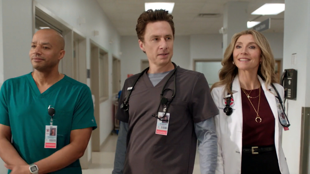 Drei Personen in Arztkleidung stehen in einem Krankenhausflur: Ein Mann in grünen Scrubs, ein Mann in grauer Arbeitskleidung und eine Frau im weissen Kittel. Alle tragen Stethoskope und Ausweise. Der sterile Gang mit weissen Wänden und Deckenbeleuchtung ist typisch für eine Klinik-Fernsehserie.