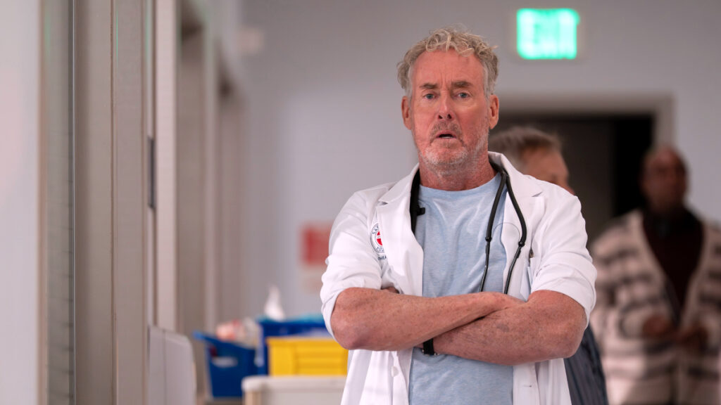 Älterer Mann mit grauen Locken und Bart in weissem Arztkittel und Stethoskop steht mit verschränkten Armen in einem Krankenhausflur. Im Hintergrund sind weitere Personen und ein grünes Ausgangsschild zu sehen.