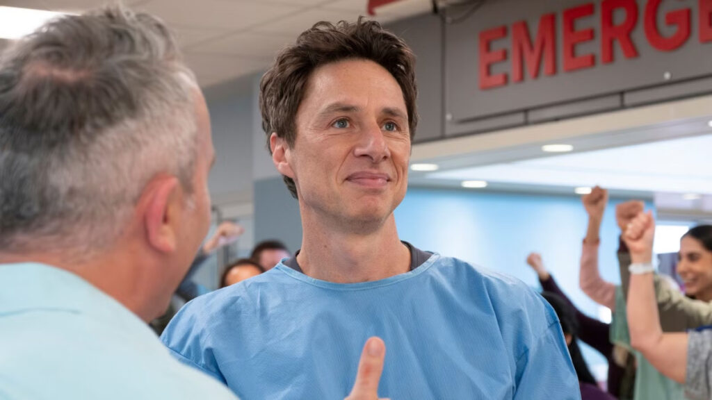 Ein Arzt in blauer Klinikuniform, vor ihm steht ein älterer Kollege. Im Hintergrund sind weitere Personen zu sehen, die jubeln, und ein «EMERGENCY»-Schild ist sichtbar. 