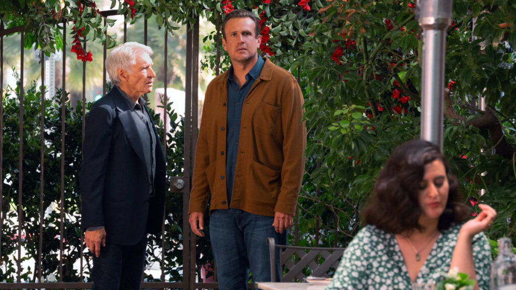 Zwei Männer stehen in einem Garten vor einem schmiedeeisernen Zaun mit üppigem Grün und roten Beeren. Links ein älterer Mann mit weissem Haar in dunkler Jacke, rechts ein jüngerer in brauner Jacke. Im Vordergrund sitzt eine Frau mit dunklen Haaren an einem Tisch.