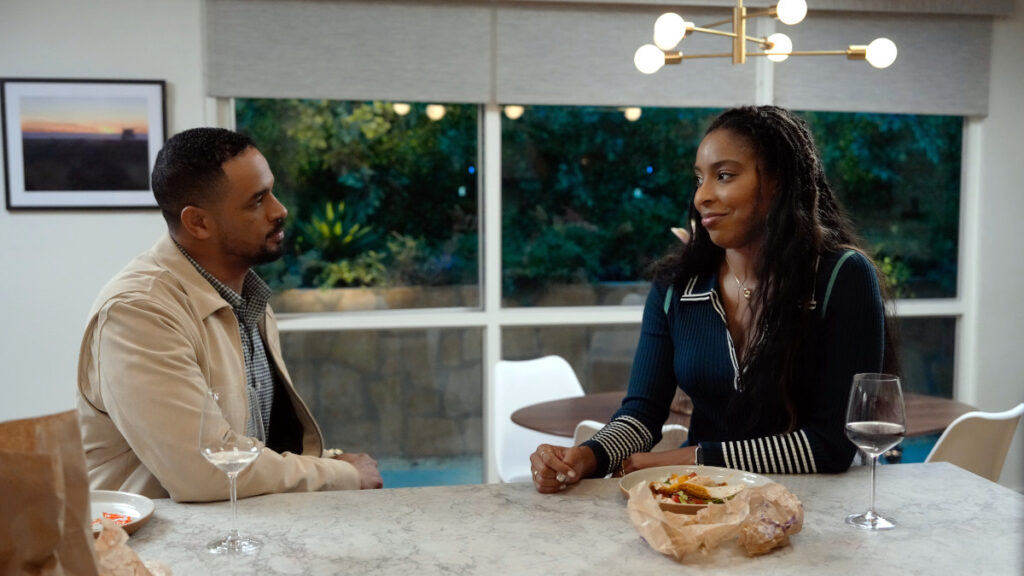 Ein Mann und eine Frau sitzen an einem Marmortisch in einer modernen Küche und unterhalten sich. Vor ihnen stehen Weingläser und Essen auf dem Tisch. Im Hintergrund sind grosse Fenster mit Blick ins Grüne und eine stilvolle Pendelleuchte zu sehen.