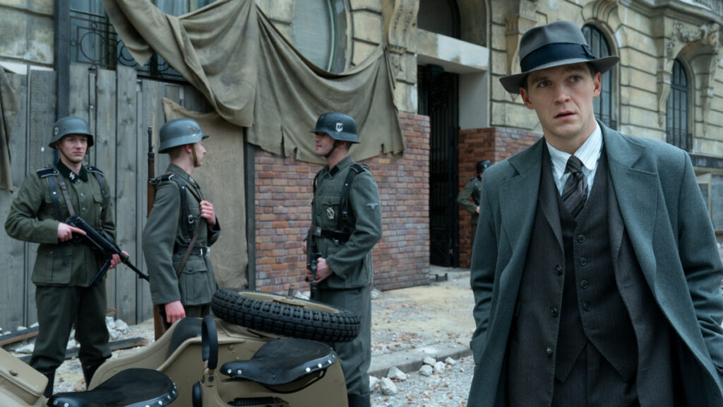 Ein Mann in grauem Anzug und Hut steht vor einem beschädigten Gebäude. Im Hintergrund sind mehrere Soldaten in grünen Uniformen und Helmen mit Gewehren zu sehen. Die Szene wirkt wie aus einem historischen Drama über den Zweiten Weltkrieg.