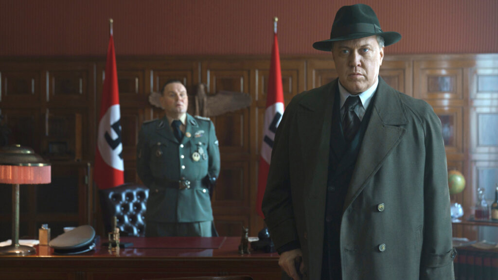 Ein Mann in dunklem Mantel und Hut steht im Vordergrund eines holzgetäfelten Büros. Im Hintergrund ein Offizier in grüner Uniform vor zwei roten Fahnen mit weissen Kreisen und schwarzen Symbolen. Die Szene wirkt wie aus einem historischen Drama der 1940er-Jahre.