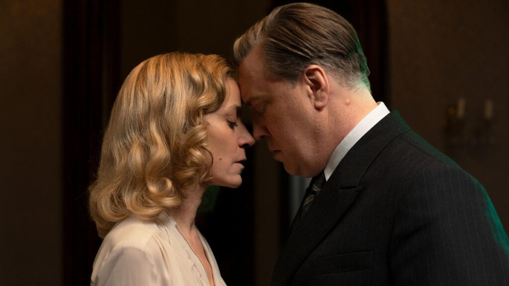 Zwei Personen in einer intimen, romantischen Pose mit den Stirnen aneinander. Die Frau trägt blonde Locken und ein helles Oberteil, der Mann einen dunklen Anzug mit Krawatte. Die Szene wirkt emotional und dramatisch.