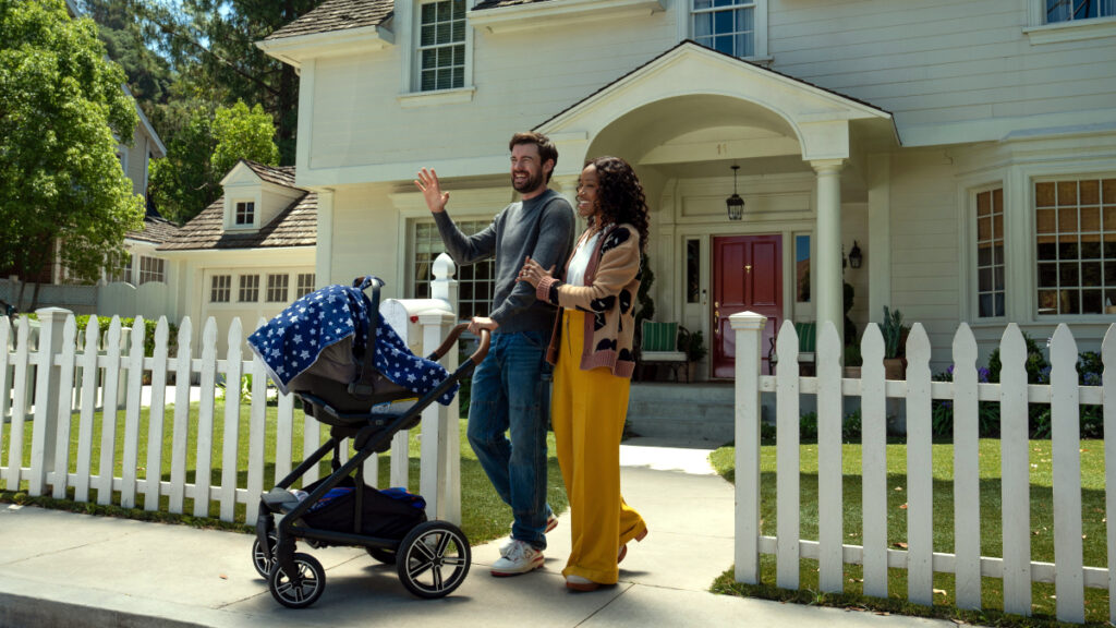 Ein Paar mit Kinderwagen steht vor einem weissen Lattenzaun und winkt fröhlich. Im Hintergrund ist ein grosses, helles Einfamilienhaus mit überdachtem Eingang zu sehen. Die Szene wirkt wie aus einer amerikanischen Vorstadtsiedlung und vermittelt eine idyllische Familienatmosphäre.