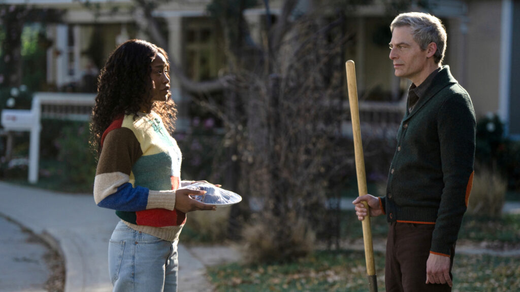 Eine junge Frau mit dunklen Locken in buntem Strickpullover hält einen Teller und unterhält sich mit einem älteren Mann mit grauen Haaren, der eine Schaufel in der Hand hält. Sie stehen auf einem Gehweg in einer Wohngegend bei Tageslicht.