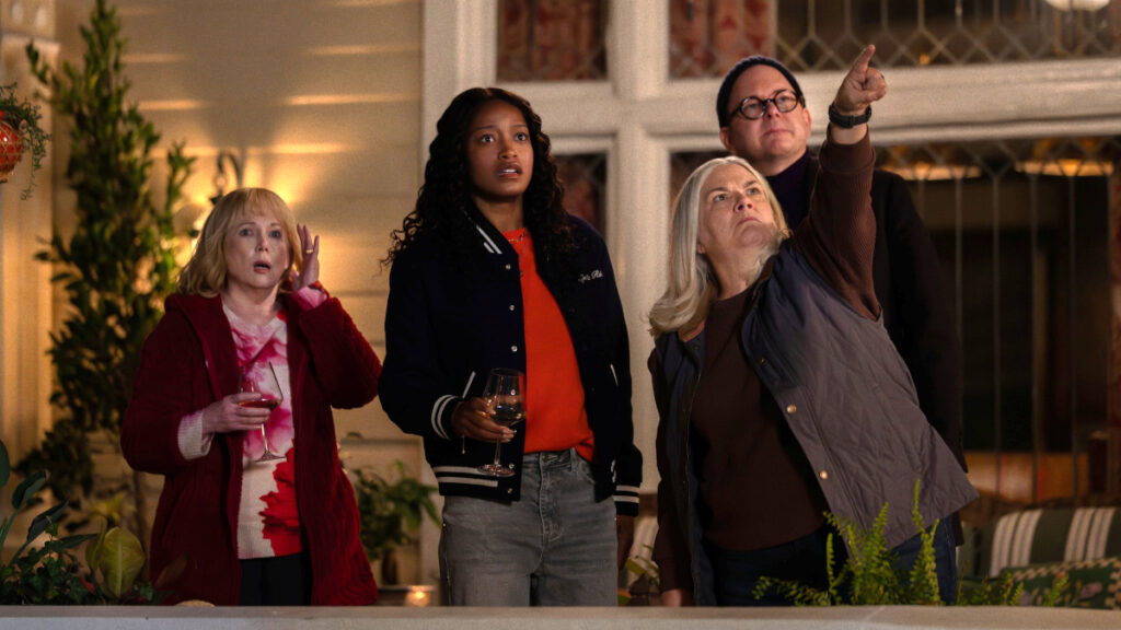 Vier Personen auf einer Veranda schauen gemeinsam leicht nach oben. Von links: blonde Frau in rotem Mantel mit Getränk, junge schwarze Frau in Bomberjacke, ältere blonde Frau und Mann mit Brille, der nach oben zeigt. Szene spielt vor einem beleuchteten Gebäude.