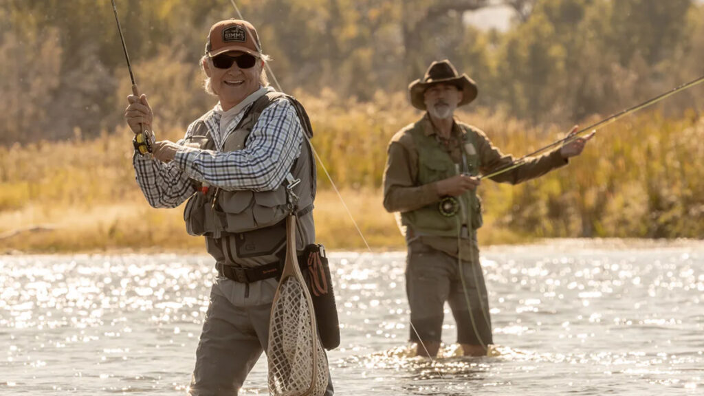 Zwei Angler stehen bei goldenem Licht in einem Fluss. Der vordere Mann trägt Sonnenbrille, Kappe und Anglerweste, hält eine Rute und ein Netz. Im Hintergrund steht ein zweiter Angler mit Hut. Herbstliche Bäume säumen das Ufer.