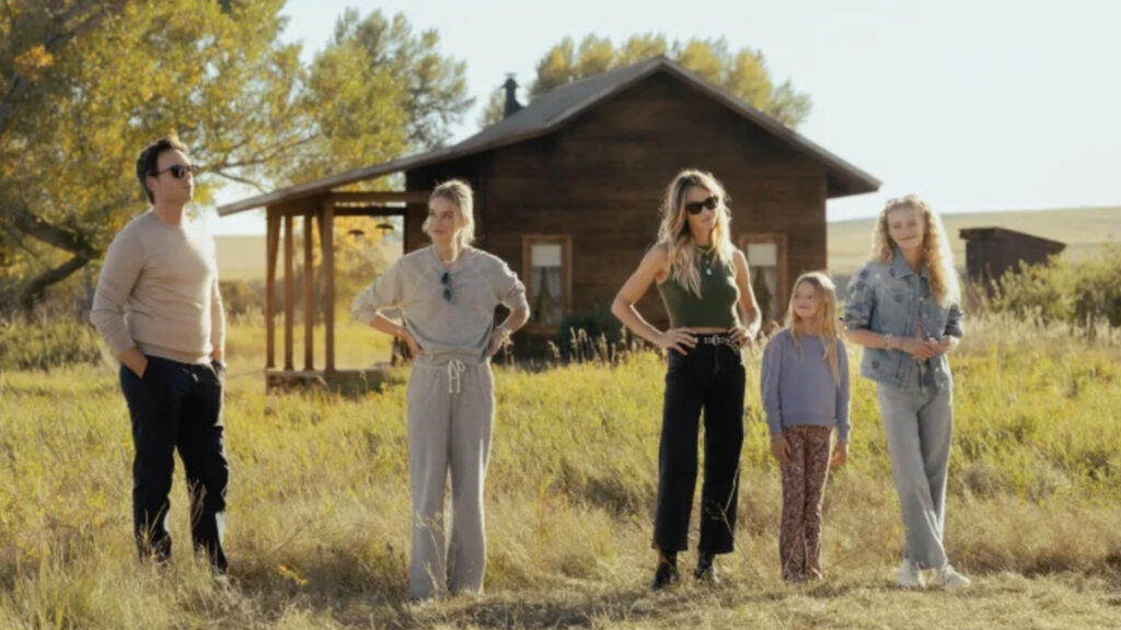 Fünf Personen stehen in einer Reihe auf einer Wiese vor einem rustikalen Holzhaus. Von links: Mann in hellem Pullover, Frau in grauem Trainingsanzug, Frau in grünem Top und schwarzer Hose, kleines Mädchen und blonde Frau in Jeansjacke. Herbstliche Landschaft mit Bäumen im Hintergrund.