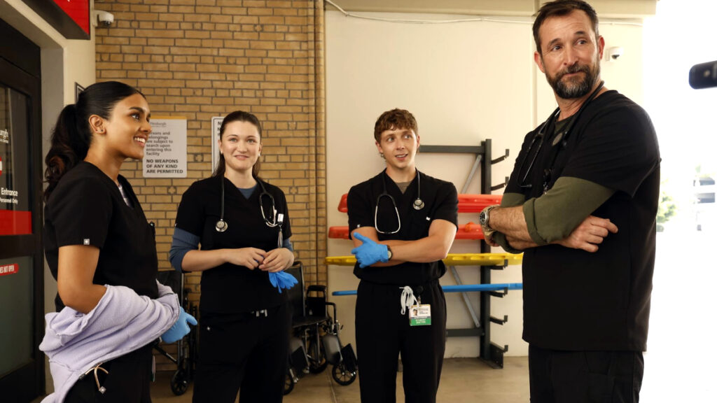 Vier Personen in medizinischer Arbeitskleidung stehen in einem Krankenhaus-Rehabilitationsraum. Drei jüngere Personen in schwarzen Uniformen mit Stethoskopen und blauen Handschuhen schauen zu einer älteren Person in schwarzem Oberteil, die mit verschränkten Armen dasteht.