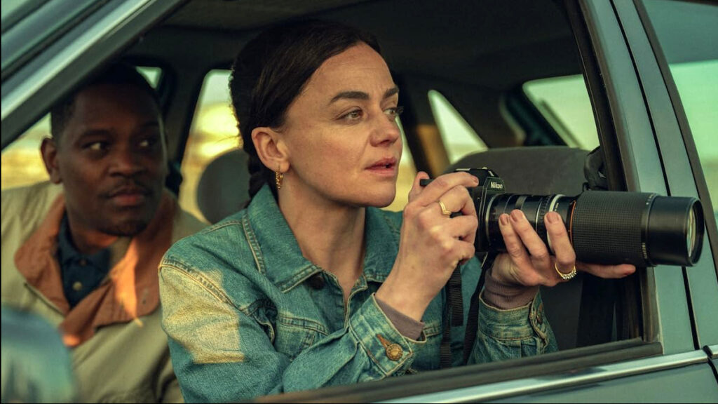 Eine Frau in Jeansjacke sitzt im Beifahrersitz eines Autos und fotografiert mit einer Nikon-Kamera durch das geöffnete Fenster. Im Hintergrund ist unscharf eine weitere Person zu sehen. Die Szene wirkt wie aus einer TV-Serie oder einem Film.