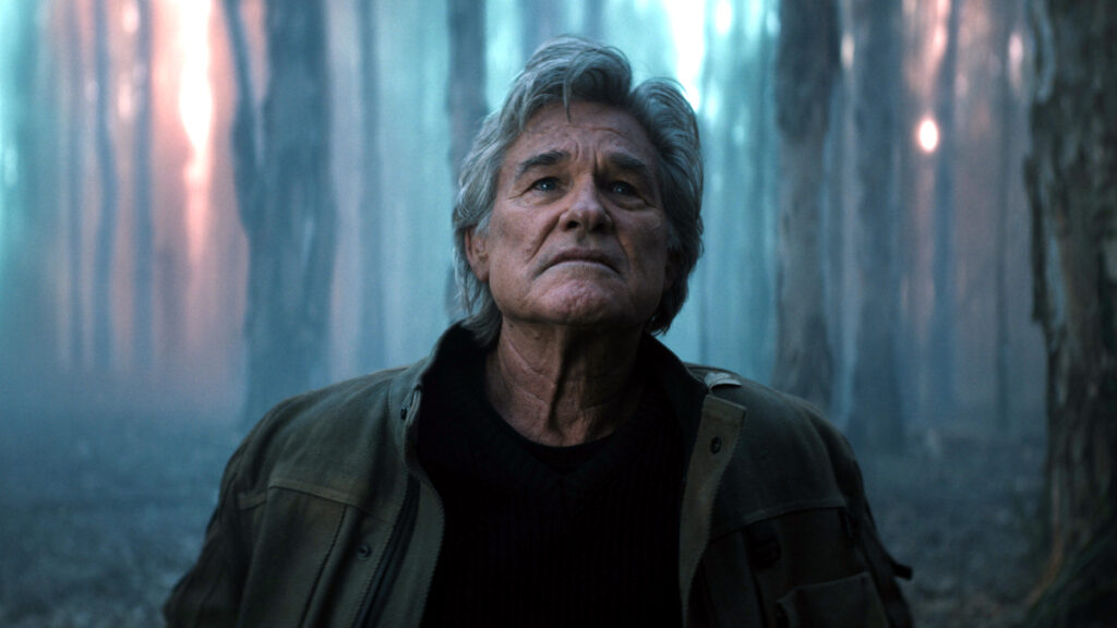 Ein älterer Mann mit grauem Haar blickt nachdenklich nach oben. Er trägt eine dunkle Jacke über einem schwarzen Shirt. Im Hintergrund sind verschwommene bläuliche und rötliche Lichter zu sehen, die eine futuristische oder Sci-Fi-ähnliche Atmosphäre schaffen.
