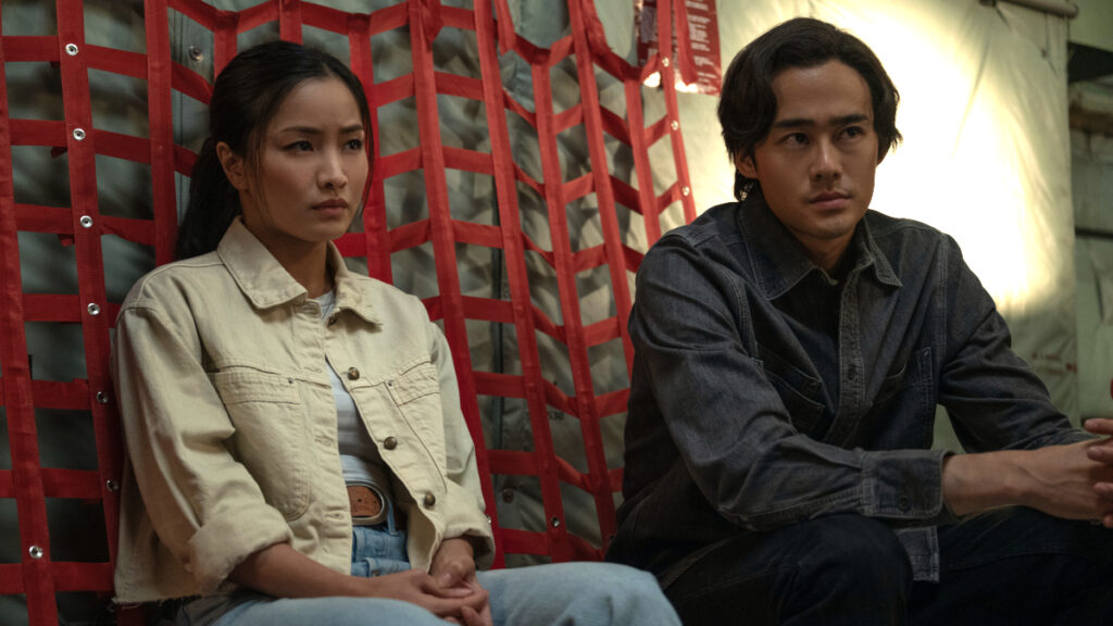Zwei asiatische Personen sitzen nebeneinander vor einem roten Netz in einem Flugzeug. Die Frau links trägt eine beige Jeansjacke und blaue Jeans, der Mann rechts ein dunkles Jeanshemd. Beide blicken nachdenklich zur Seite.