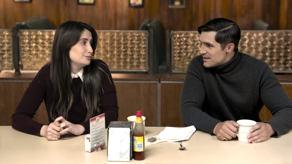 Eine junge Frau in burgunderfarbener Bluse und ein Mann in grauem Pullover sitzen sich in einem Café oder Restaurant gegenüber und unterhalten sich. Auf dem Tisch stehen eine Milchpackung, eine Flasche und eine Tasse. Im Hintergrund sind warme Holztöne und Beleuchtung zu sehen.