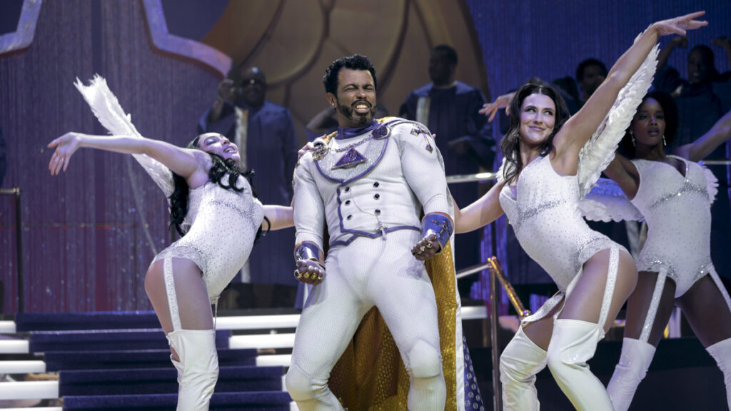 Ein Schauspieler in einem weissen Prinzenkostüm mit goldenen Details steht auf einer Theaterbühne zwischen zwei Tänzerinnen in weissen, glitzernden Outfits. Alle drei haben die Arme ausgebreitet in einer choreografierten Pose. Im Hintergrund sind weitere Ensemble-Mitglieder zu sehen.