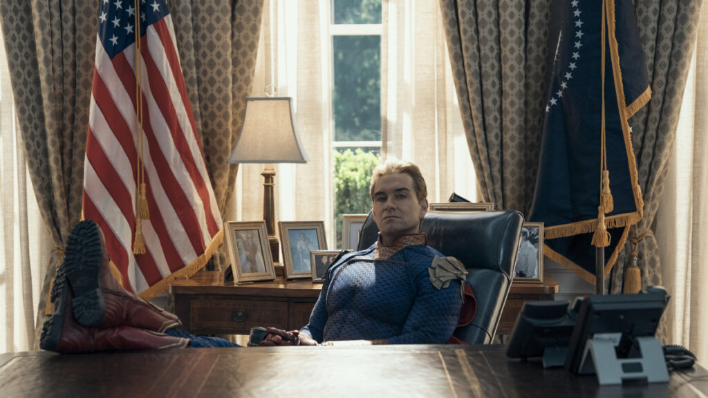 Ein Mann in einem blauen Superheldenkostüm sitzt entspannt mit hochgelegten Füssen am Schreibtisch im Oval Office. Im Hintergrund sind die amerikanische Flagge, elegant drapierte Vorhänge und gerahmte Familienfotos zu sehen.
