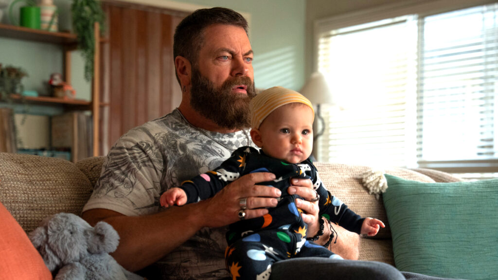 Bärtiger Mann sitzt auf beigem Sofa und hält Baby in buntem Strampler mit Dinosaurier-Motiven. Neben ihnen liegt ein grauer Plüschteddy. Im Hintergrund sind Küchenregale und ein Fenster mit Jalousien zu sehen. Gemütliche Wohnzimmerszene.