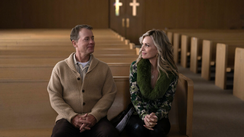 Ein Mann in beigem Strickpullover und eine Frau in grünem Schal und geblümter Bluse sitzen nebeneinander auf einer Kirchenbank. Beide schauen zur Seite und lächeln leicht. Im Hintergrund sind hölzerne Kirchenbänke und Kreuze an der Wand zu sehen.