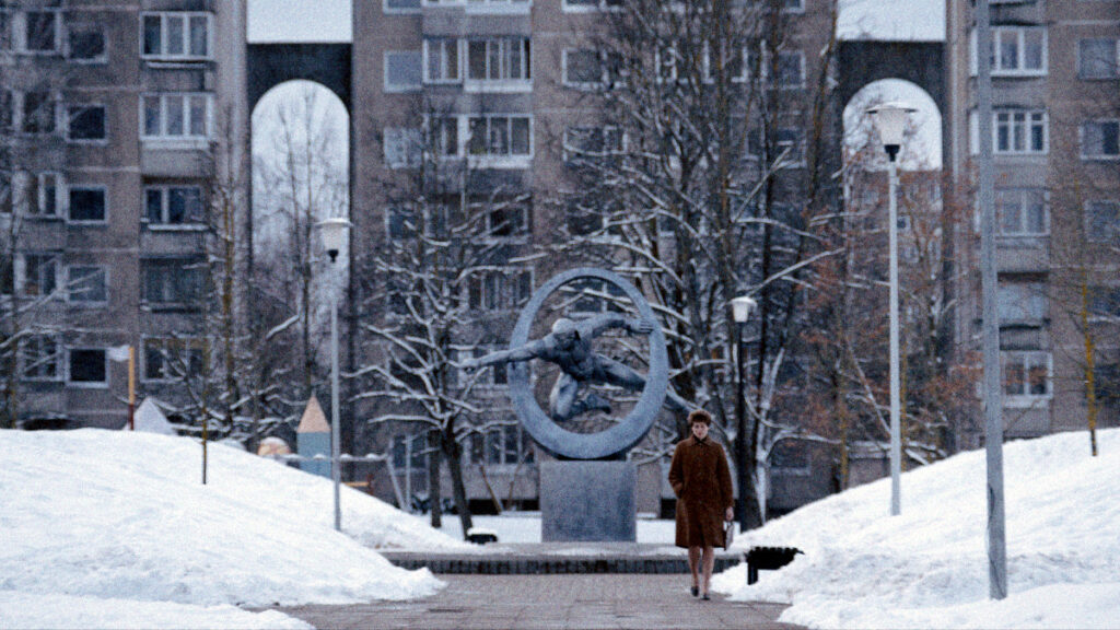 Eine Frau in braunem Mantel spaziert im Winter auf einem Weg durch einen verschneiten sowjetischen Platz. Im Hintergrund ein Sportler-Denkmal mit Ringmotiv und ein grauer Plattenbau.