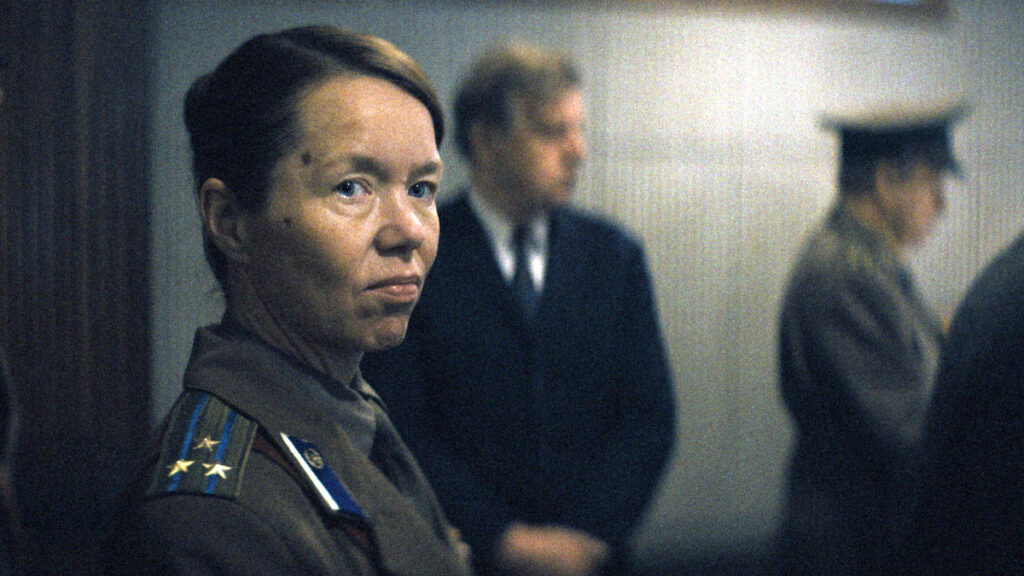 Eine Frau in sowjetischer Militäruniform mit Schulterstücken blickt ernst in die Kamera. Im unscharfen Hintergrund stehen zwei weitere Personen, eine davon ebenfalls in Uniform.