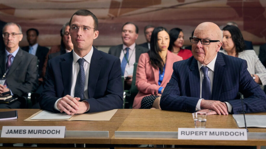 Zwei Männer in Anzügen sitzen an einem Tisch mit den Namensschildern «James Murdoch» und «Rupert Murdoch». Im Hintergrund sitzen weitere Personen. Die Szene wirkt wie eine offizielle Anhörung.