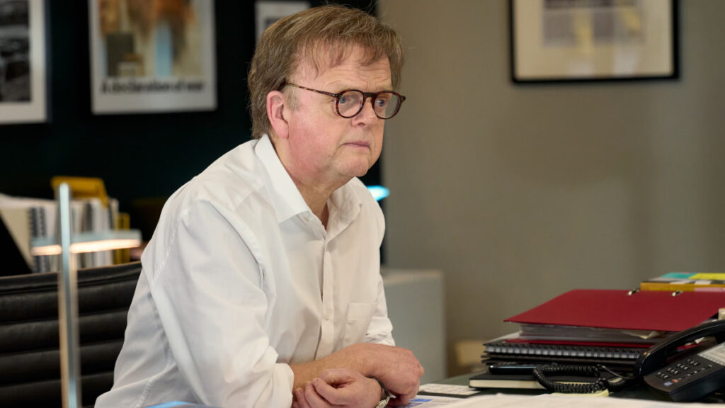 Ein älterer Mann mit Brille und weissem Hemd sitzt an einem Schreibtisch mit Akten und einem Telefon. Im Hintergrund hängen gerahmte Bilder an der Wand. 