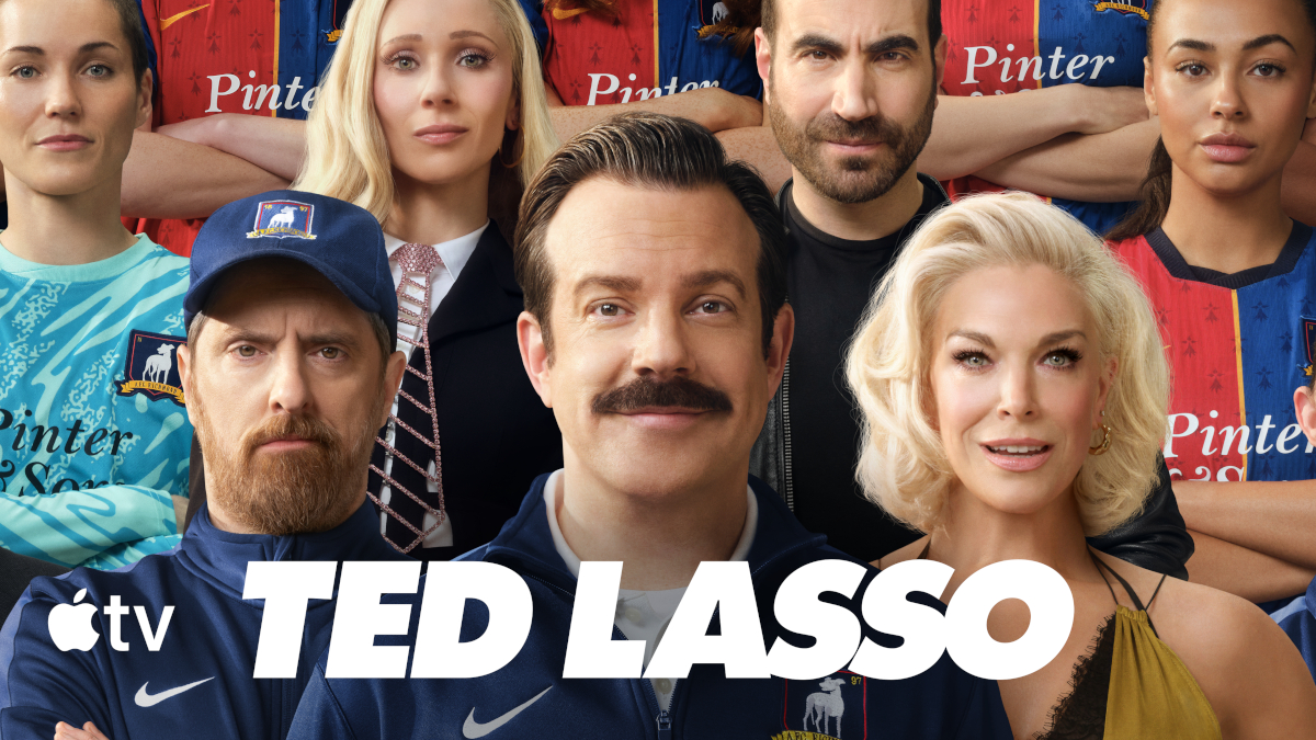 **Alt-Text:** Poster der Apple-TV-Serie «Ted Lasso»: Sieben Personen posieren vor blauen und roten Fussballtrikots. Im Zentrum ein Mann mit Schnauzbart und blauer Trainingsjacke. Unten der Serientitel «Ted Lasso».